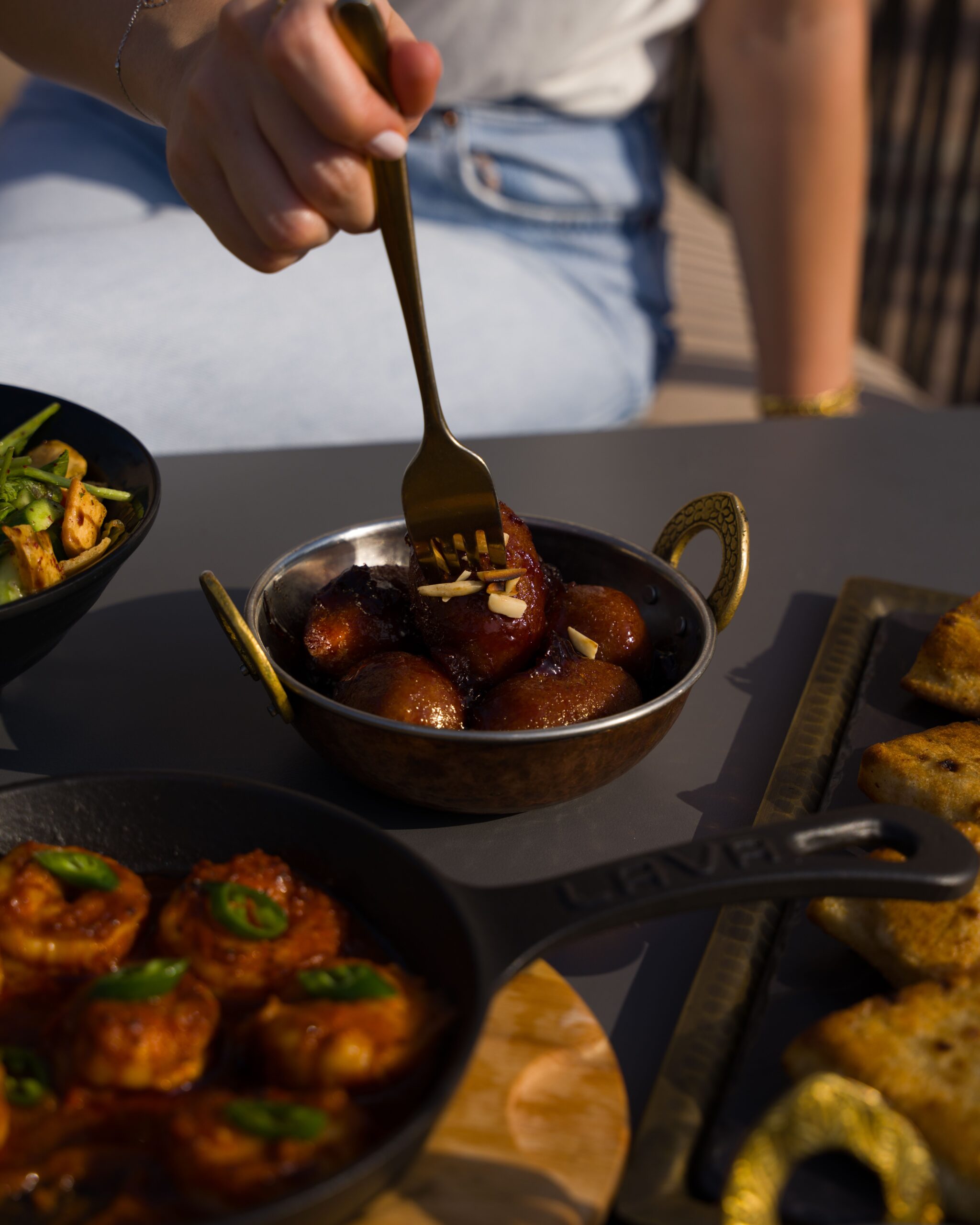 Close-up of Gulab Jamun being picked with a fork, surrounded by Indian dishes.